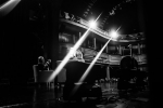 fot. Dorota Awiorko / [Ujęcie z głębi sceny, zza pleców kobiety i mężczyzny siedzących w fotelach. W tle widownia teatru składająca się z trzech poziomów. U góry rozbłyski dwóch reflektorów.]