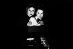 fot. Dorota Awiorko / [Czarno-białe zdjęcie dwóch przestraszonych kobiet stojących za uśmiechniętym mężczyzną siedzącym bokiem do kobiet. Światło pada tylko na twarze, wokół mrok.]