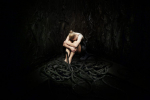 fot. Dorota Awiorko / [Kobieta w krótkiej czarnej sukience siedząca na podłożu z podkulonymi nogami, które oplata rękami opierając głowę na kolanach. W tle czarna ściana o nierównomiernej fakturze, przed kobietą kłębowisko grubych, czarnych lin.]