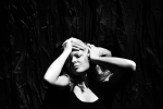 fot. Dorota Awiorko / [Kobieta trzymająca swoje dłonie na obcych dłoniach znajdujących się na jej głowie, wyłaniających się zza znajdującej się za nią czarnej kotary. Kobieta ubrana w krótką czarną sukienką na ramiączkach.]