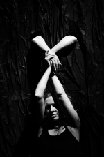 fot. Dorota Awiorko / [Kobieta trzymana za wyciągnięte w górę, złączone ręce przez inne ręce wyłaniające się ponad nią zza ciemnej kotary.]