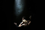 fot. Dorota Awiorko / [Mężczyzna w garniturze opierający się o ścianę pokrytą nierównościami, zwrócony w jej stronę i kobieta z gołymi ramionami przytulająca się do pleców mężczyzny. Tło ciemne.]