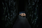 fot. Dorota Awiorko / [Korytarz pomiędzy ścianami utworzonymi z intensywnie pozaginanego, ciemnego tworzywa. W głębi, na końcu korytarza na podłożu leży kobieta w pozie embrionalnej.]