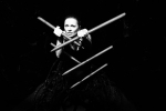 fot. Dorota Awiorko / [Patrząca prosto w obiektyw kobieta trzymająca, krzyżujące się wystające ze ścian, więżące ją w rogu długie, zaostrzone kołki. Tło czarne.]