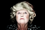 fot. Dorota Awiorko / [Zbliżenie na twarz kobiety z miną pełną powagi, z szeroko otwartymi, niebieskimi oczami i peruką z kręconymi włosami. Tło ciemne.]