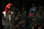 fot. Dorota Awiorko / [Po lewej postać w białym garniturze z czerwoną maską ptaka z wielkim dziobem. Resztę zdjęcia zajmuje płyta pleksi, w której odbijają się i rozmazują w jej nierównościach kolorowe, niedookreślone kształty.]