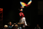 fot. Dorota Awiorko / [W centrum postać w białym garniturze z wielką, czerwoną maską ptaka, wokół postaci skupiona gromadka dzieci. Powyżej równomiernie rozmieszczone, zawieszone w przestrzeni kolorowe, zabawkowe ptaki. Tło czarne.]