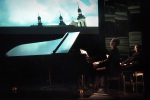 fot. Dorota Awiorko / [Pianista przy fortepianie odbijający się w stojącym za nim lustrze. Powyżej ekran ze zdjęciem wieżyczek budynków na starym mieście na tle błękitnego nieba z białymi chmurami.]